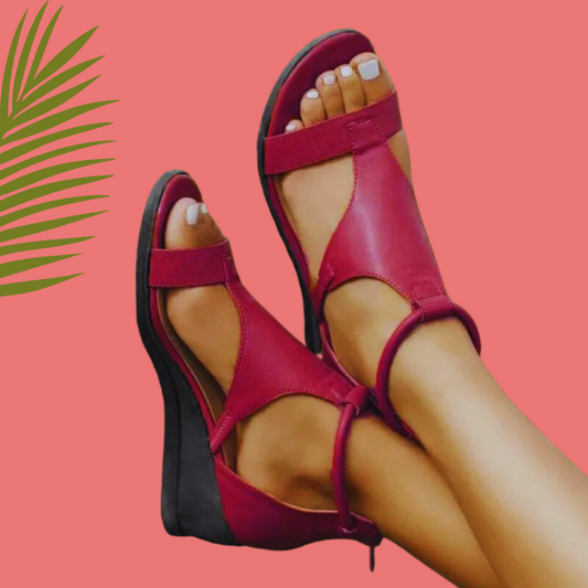 A pair of red orthopedic leather sandals with a thick sole and ankle straps, displayed against a pink background with a palm leaf motif.